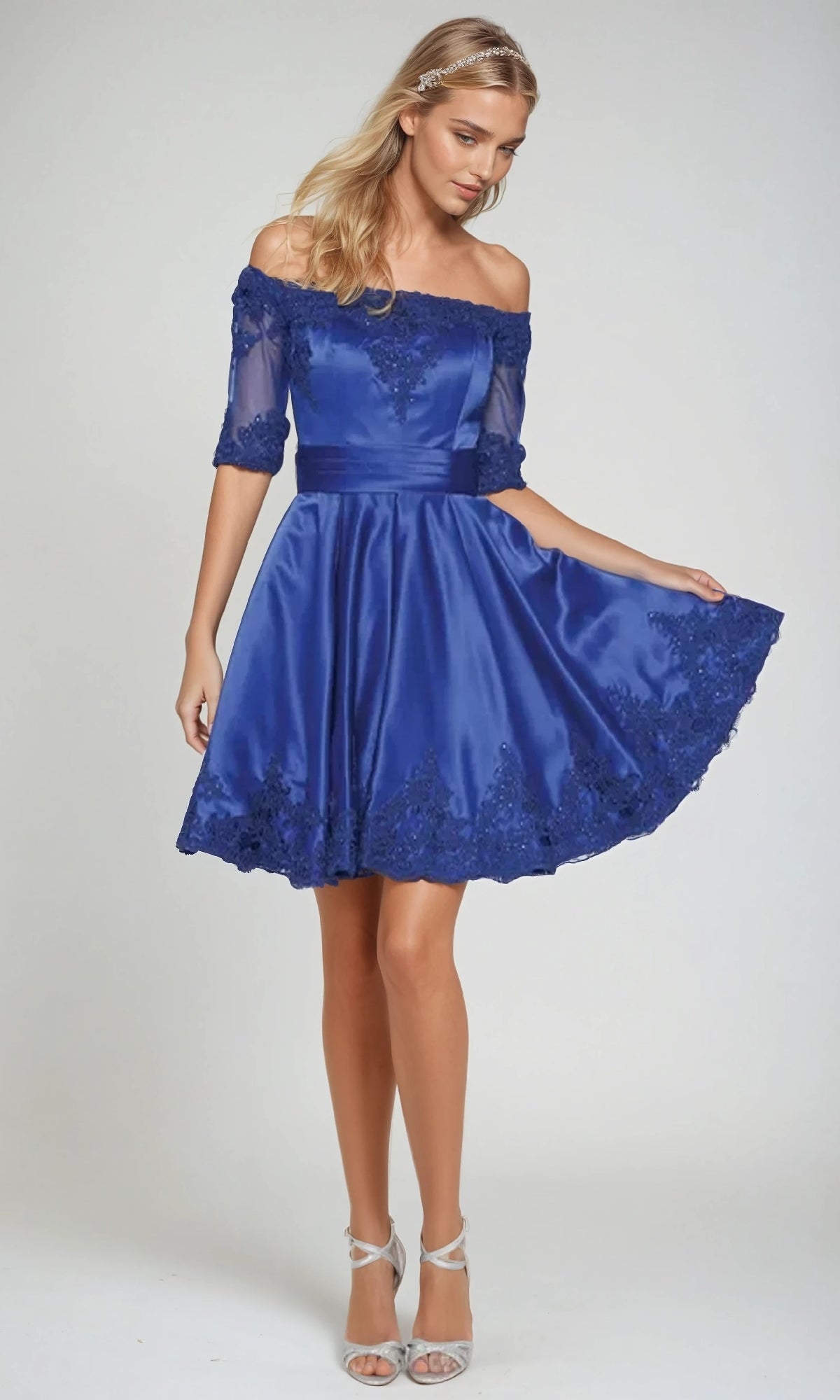 A woman models the Juliet 804, a royal blue, knee-length, off-the-shoulder semi-formal dress with lace trim, posing against a plain background and holding out one side of the skirt.
